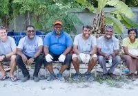 Parte da equipe do projeto do Soneva para redução do uso de plástico nas Maldivas. Cinco homens e uma mulher sentam-se lado a lado, com pé na areia e fundo de vegetação verde.
