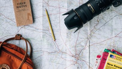 planejar-uma-viagem-agora Planejar uma viagem: mapa, câmera fotográfica, mochila e cadernos estão sobre uma mesa.