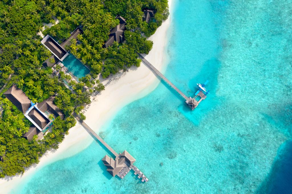 Foto aérea de hotel nas ilhas Maldivas. Água azul turquesa do mar divide a imagem com uma faixa de areia branco e um pedaço de terreno preenchido por vegetação. Staycation.