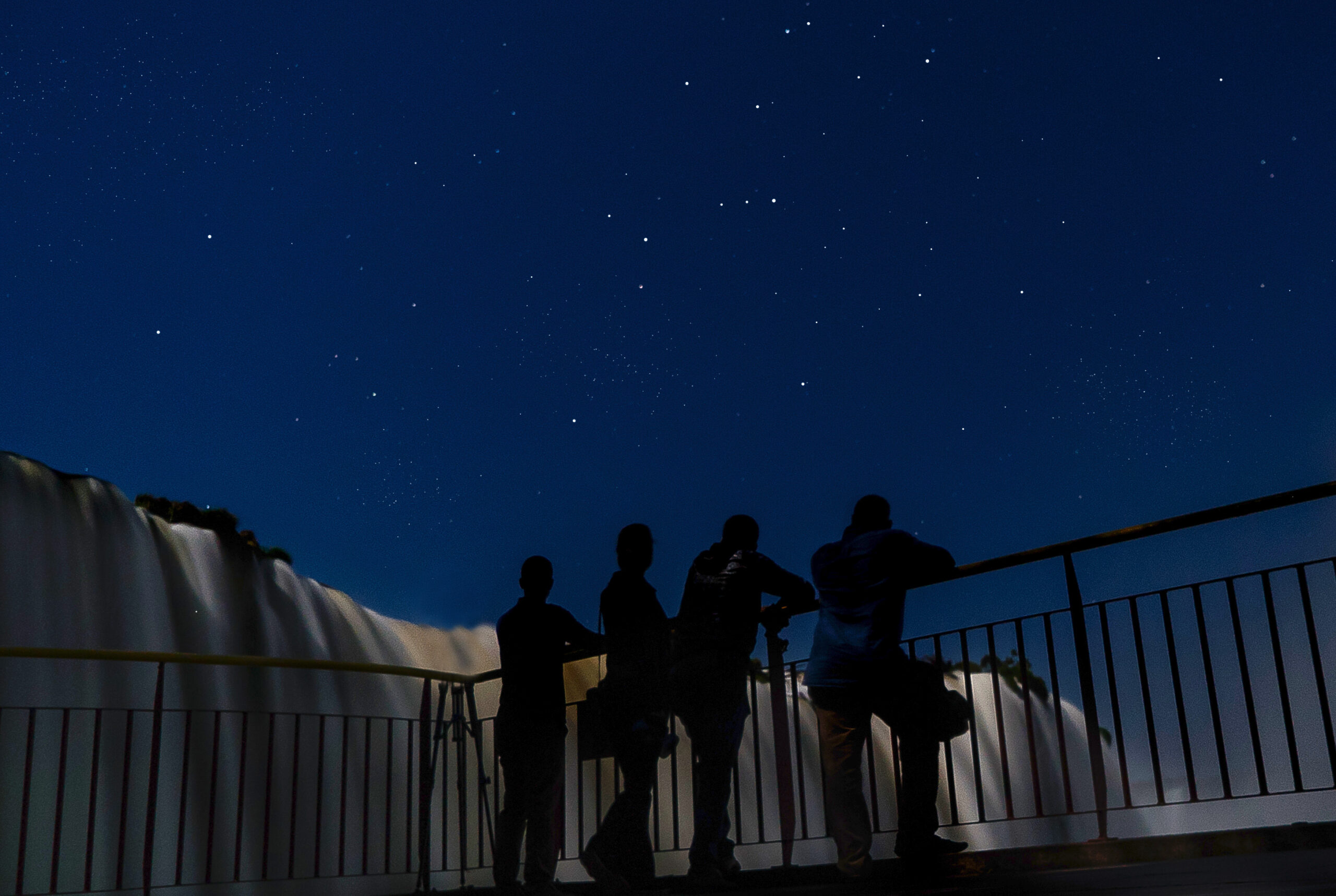 Quatro pessoas estão de costas para a foto, apoiadas em uma cerca que a passarela possui. Estão observando as quedas d'água das Cataratas do Iguaçu, que preenchem o fundo da paisagem junto com um céu estrelado.