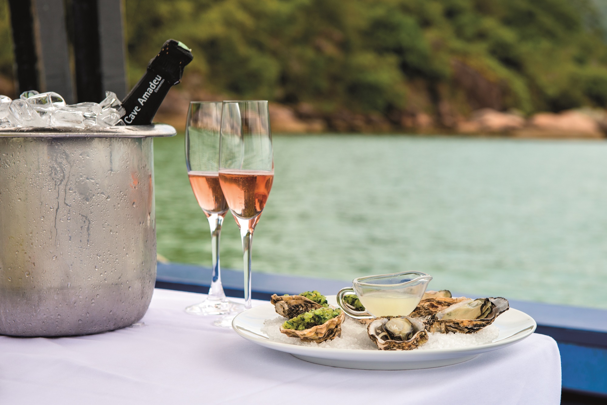 Foto mostra mesa posta dentro de barco com toalha branca, taças de champagne e um prato com ostras.