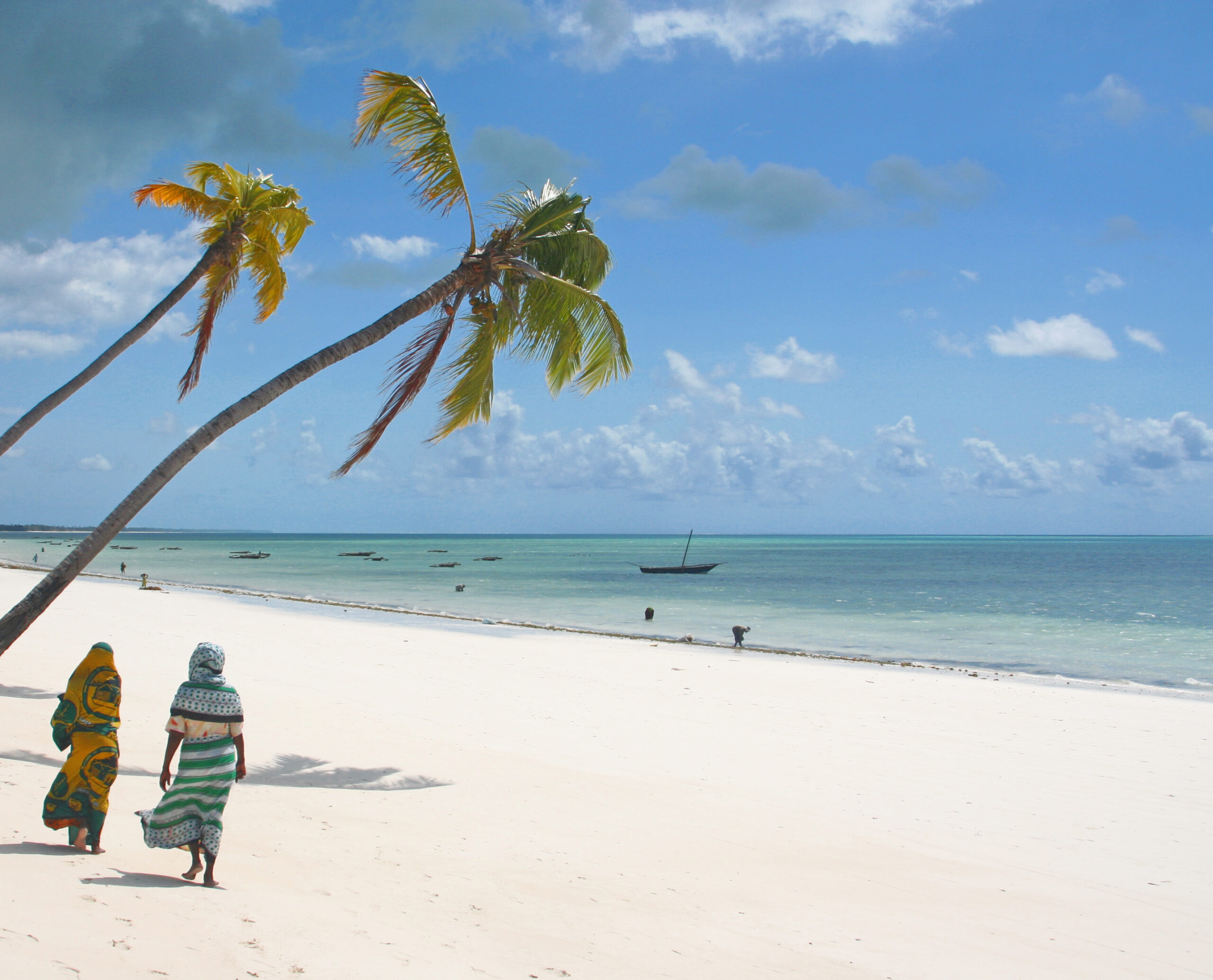 Praia na ilha de Zanzibar, na Tanzânia, com areia clara, mar azul e dois coqueiros inclinados. Há duas moças com trajes muçulmanos caminhando na areia. Boa combinação de destino com Serengeti safari.