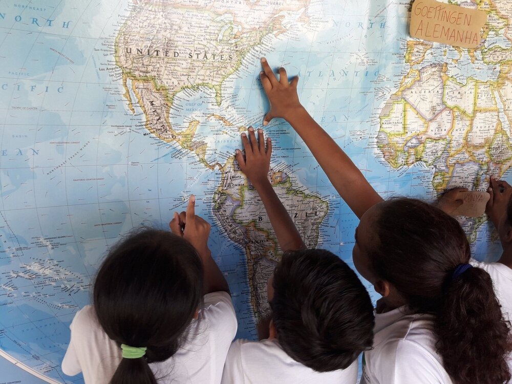 Crianças da escola na Serra do Amolar estão de costas e apontam para um ponto no mapa mundi que fica na parede.