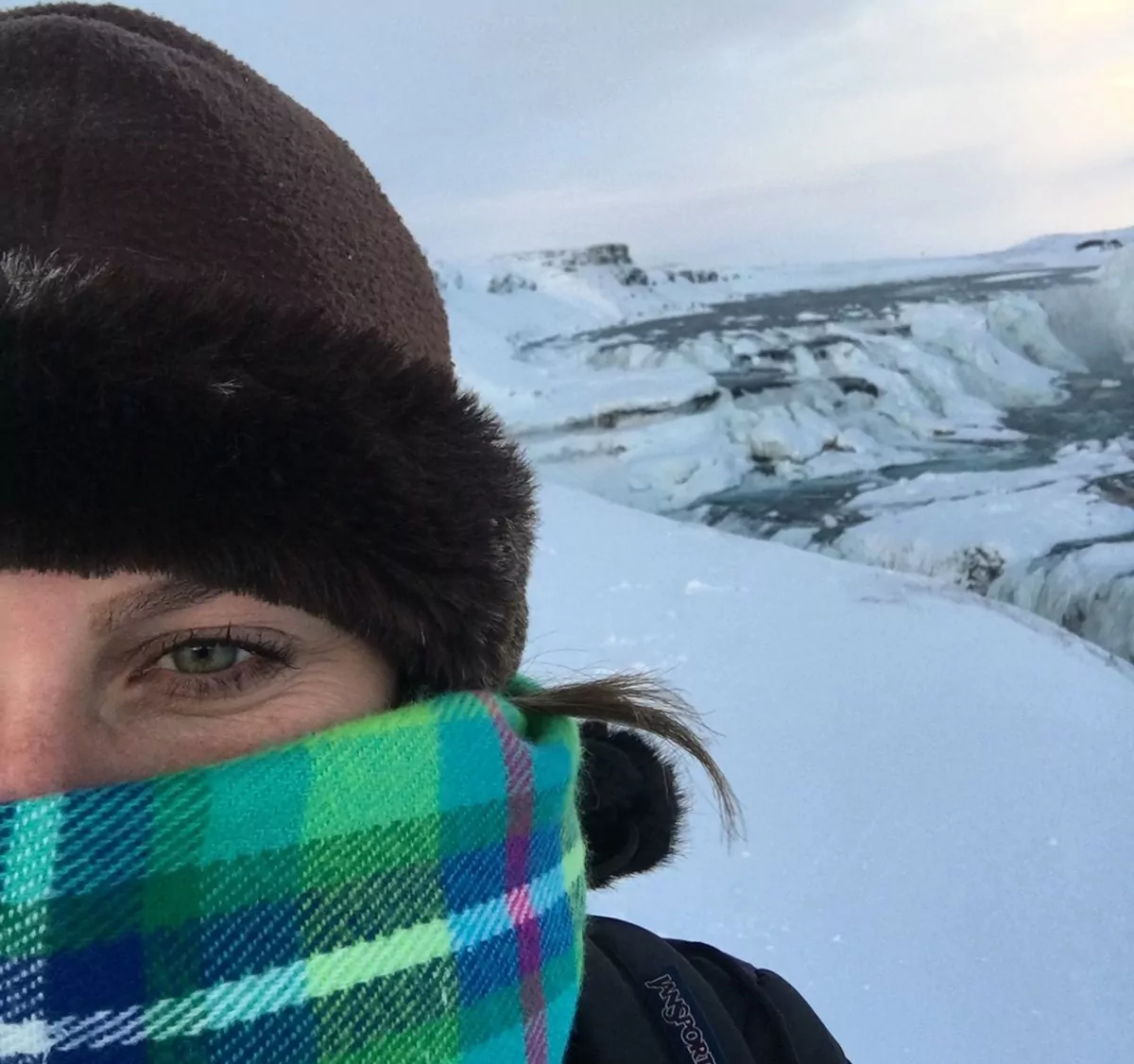 Tatiana de capuz e cachecol mostra ao fundo como é a Islândia, coberta de neve.