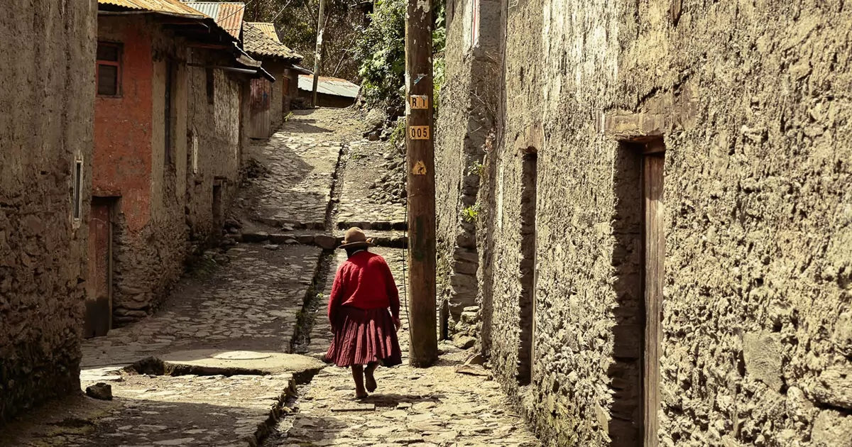 Mulher peruana com roupa vermelha caminha por uma rua de pedras, com casas de pedras. Tendências de viagem em 2022 com agências de viagem.