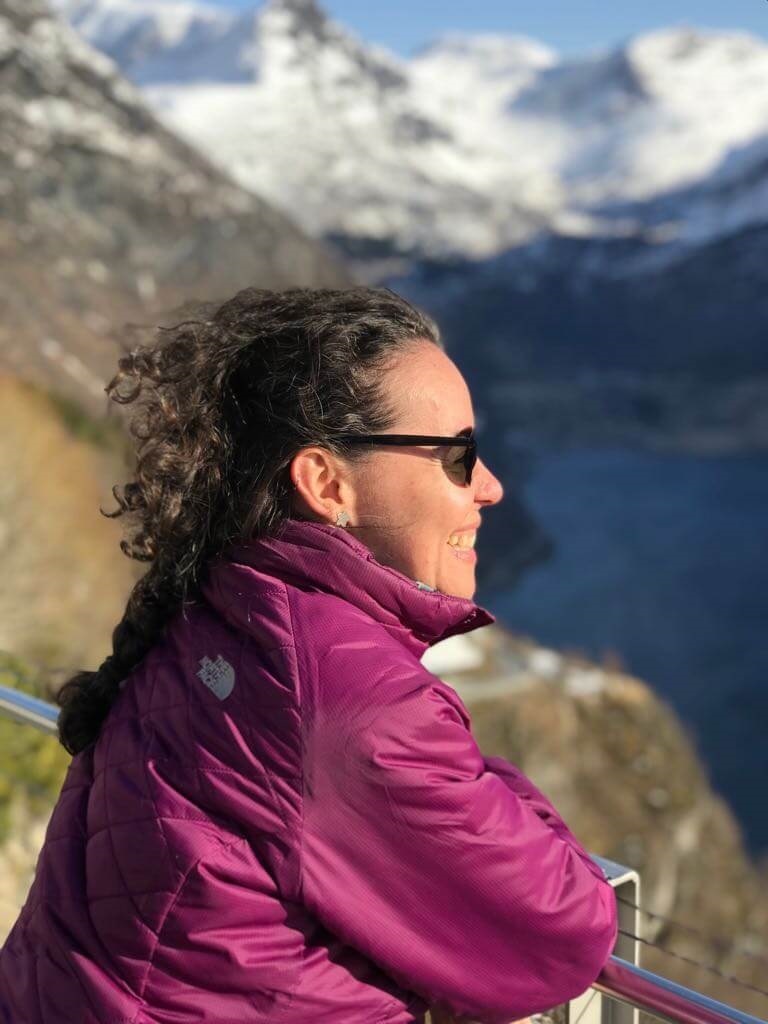 Mulher de posa de lado, apoiada em uma grade baixa, sorrindo e olhando para o horizonte a sua frente. Ao fundo, os fiordes noruegueses, formados entre montanhas.