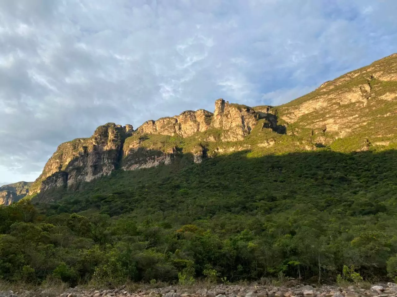 Montanha se ergue no Vale do Pati. O sol reflete sobre a formação rochosa, enquanto o céu está um pouco nublado e abaixo há vegetação verde.