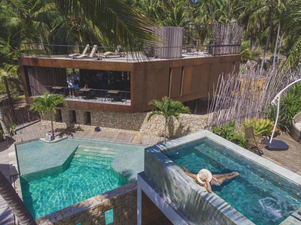 Hotel Pedras do Patacho, na Praia do Patacho, Alagoas. Uma construção em meio à vegetação, com uma piscina no centro, em que uma mulher de chapéu toma sol.