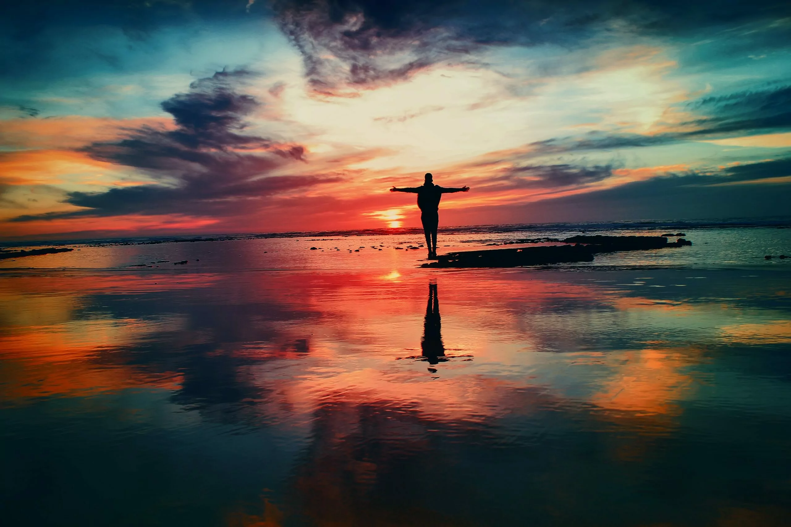 Pessoa na praia, frente para o mar, braços abertos em forma de cruz observando o por do sol em viagem espiritual