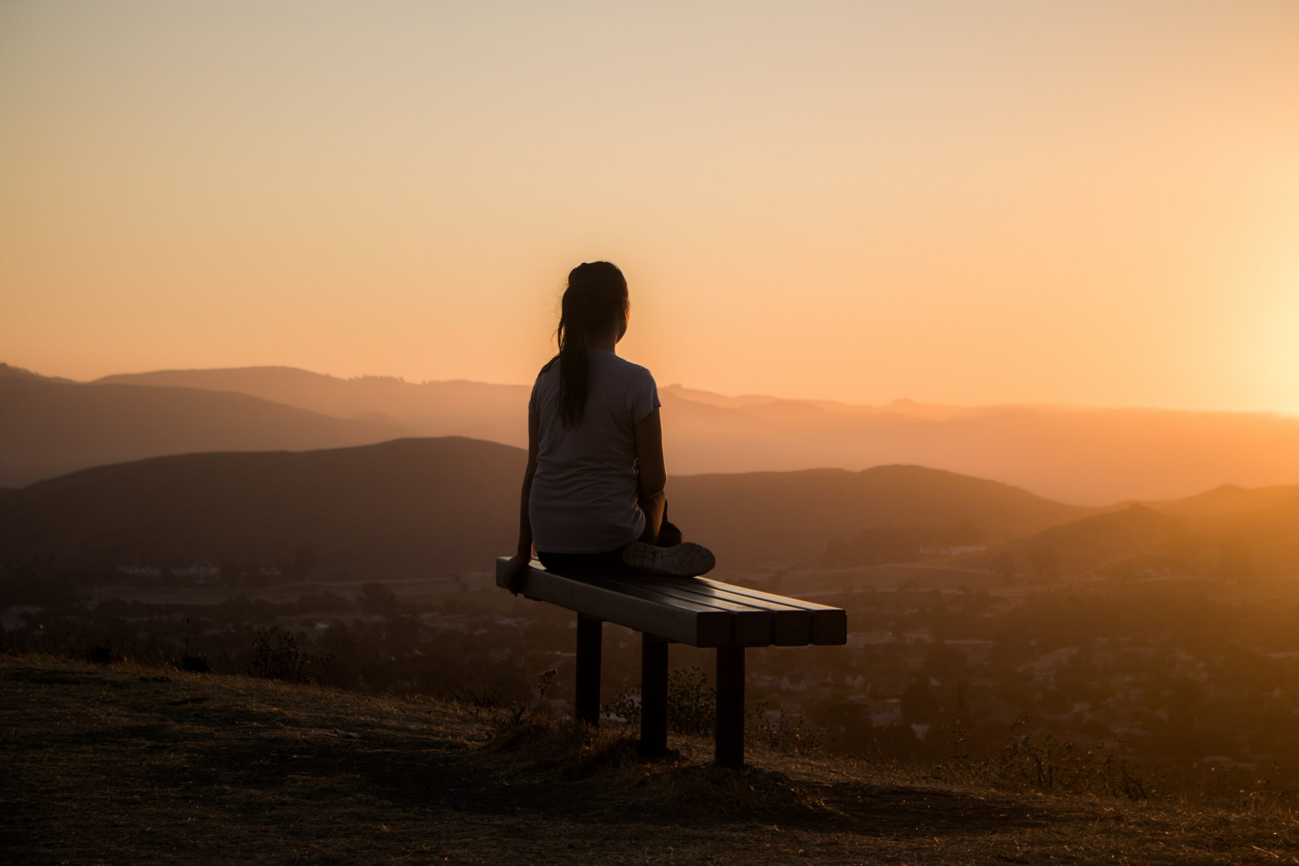 imagem mostra mulher sentada em um banco de madeira, ao por do sol, no topo de uma montanha, aparentemente meditando