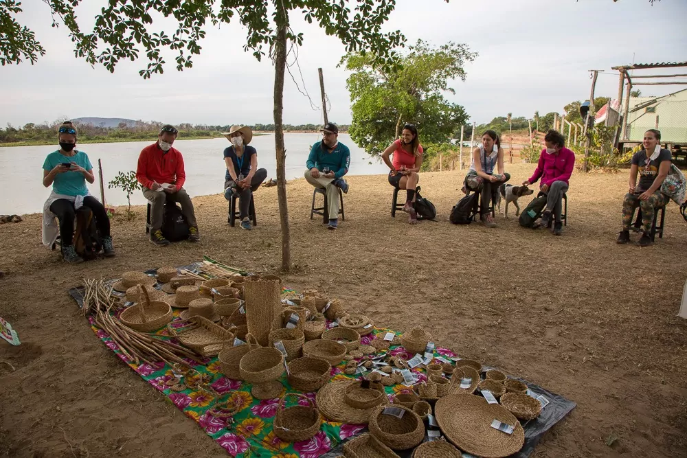 turistas sentados ao redor de esteira, à beira de um rio, estudem e discutem artesanato produzido por povos originários