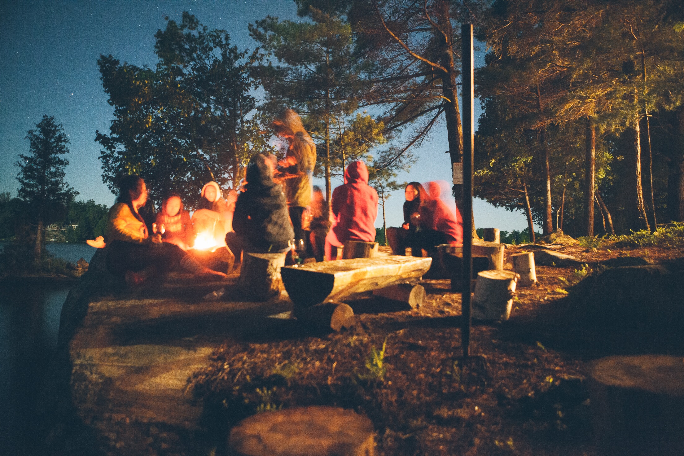 família ao redor de fogueira ao anoitecer sentados em bancos rústicos de madeira à beira de rio e árvores ao fundo